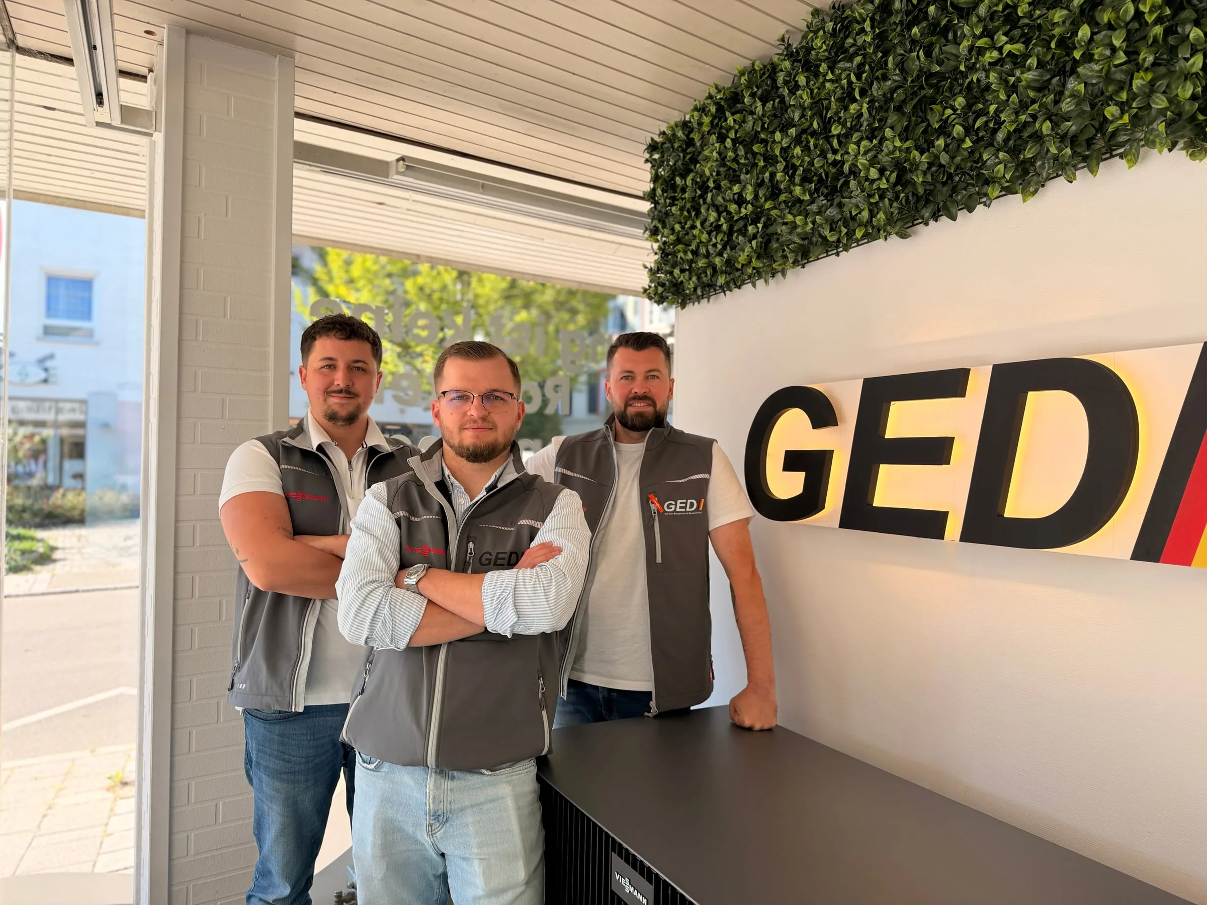 Drei Mitarbeiter des GED Teams in grauen Firmenwesten im Showroom in Trossingen neben dem GED Logo